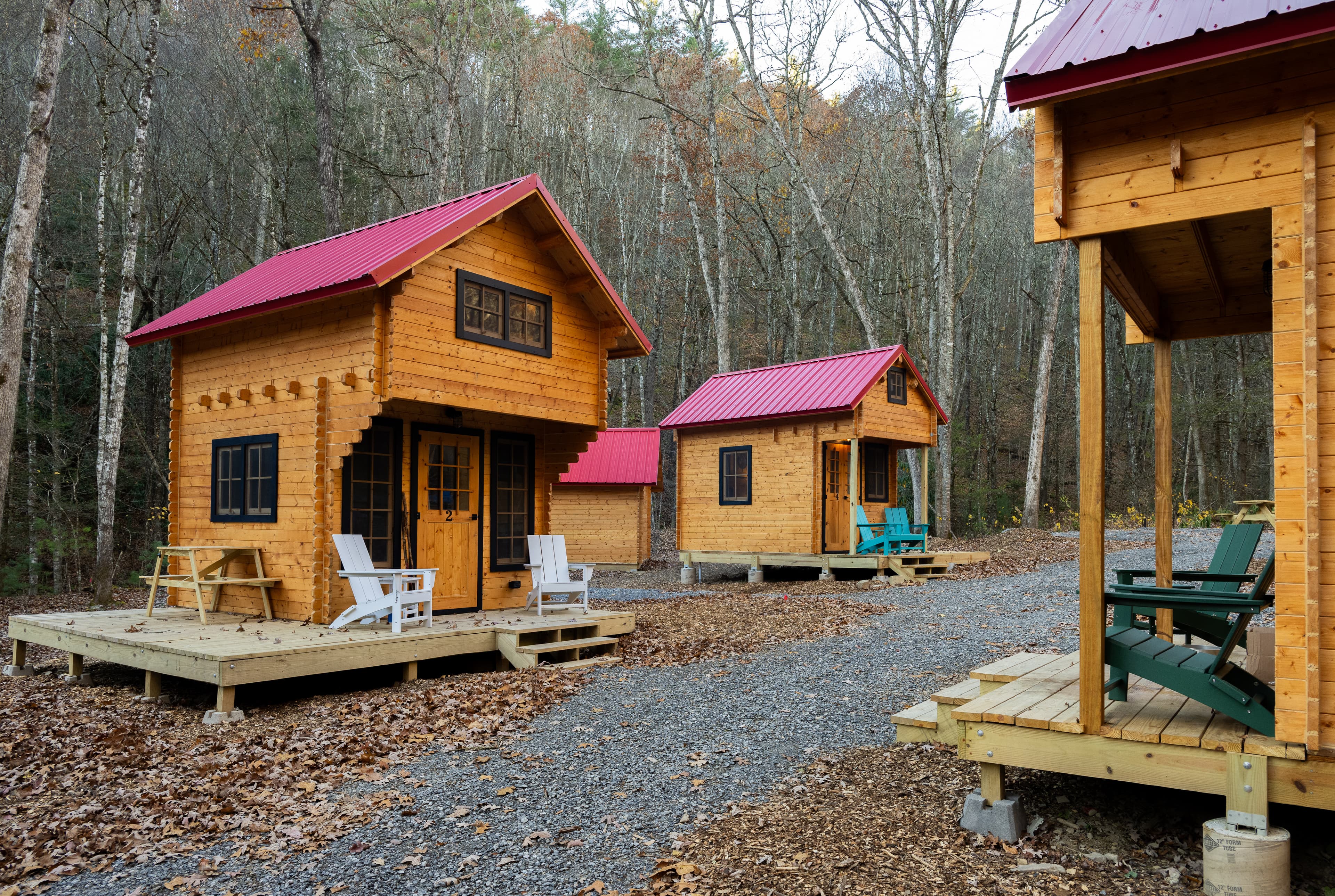 Three wooden cabins with red roofs nestled in a forest, surrounded by gravel paths and autumn leaves.