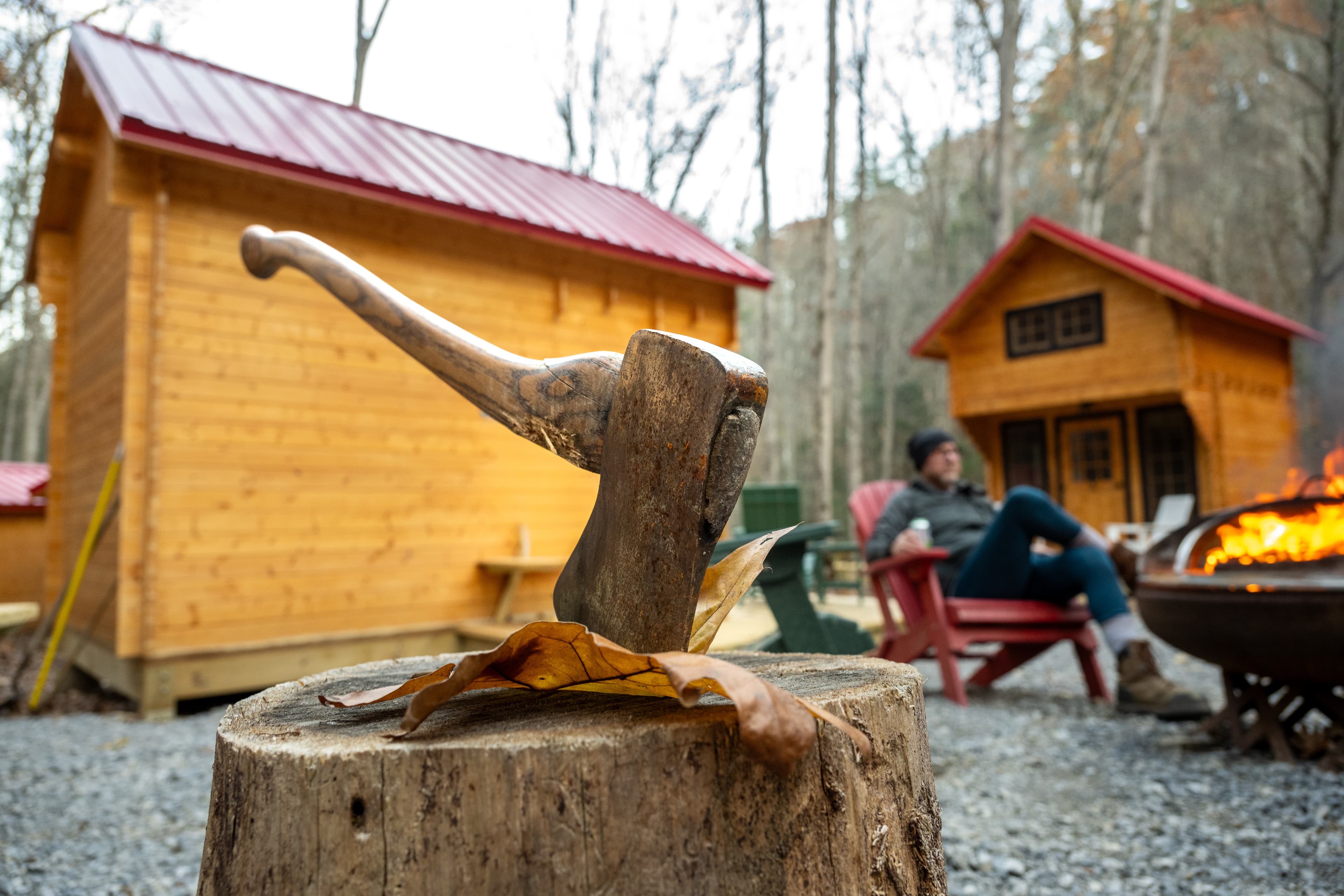 An axe rests on a wooden stump in front of two cabins, with a person sitting nearby and a fire pit in the foreground.