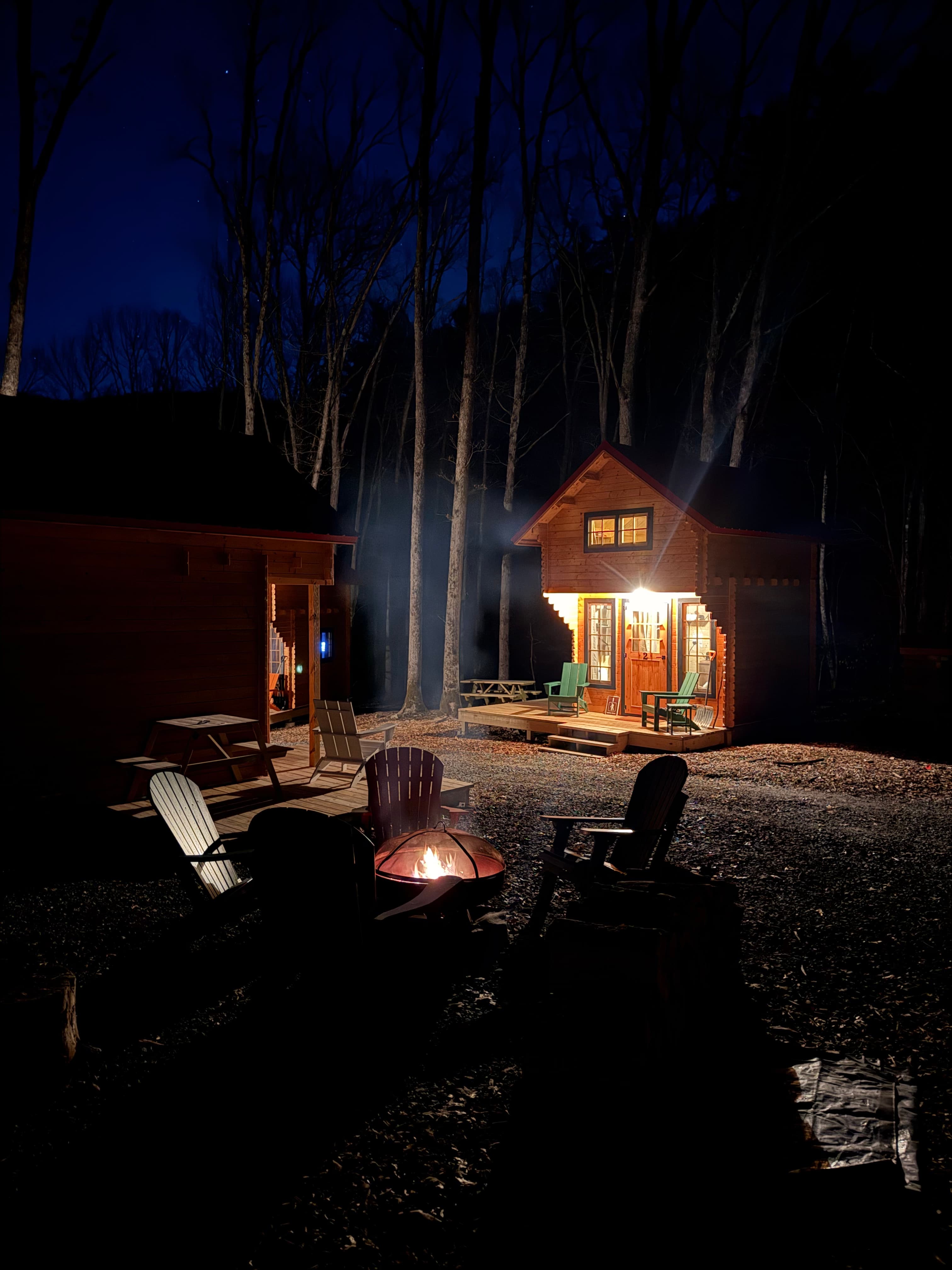 A cozy cabin illuminated at night, surrounded by trees and a glowing campfire.